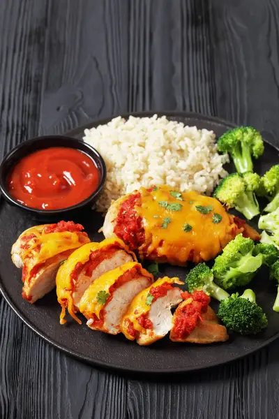 baked chicken breasts with tomato salsa, cheddar cheese and parsley on black plate with broccoli, brown rice and tomato sauce on black wooden table, vertical view, close-up
