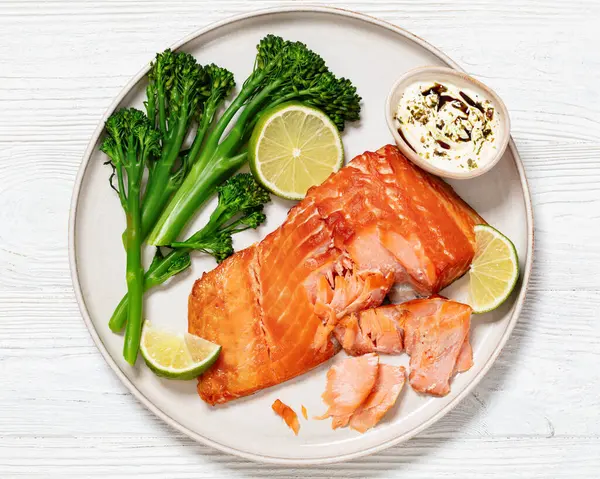 hot smoked salmon with cooked broccoli, lime and yogurt based sauce on plate on white wooden table, horizontal view from above, flat lay, close-up