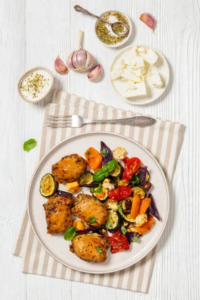 baked boneless chicken thighs with baked mixed vegetables and cubed feta cheese on plate on white wooden table with ingredients and fork, vertical view from above