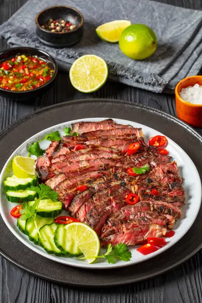 stock image crying tiger, grilled and thinly sliced ribeye steak with cucumber and lime on plate on black wooden table with spicy dipping sauce, thai cuisine, vertical view from above, close-up