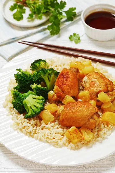 roasted boneless chicken thighs with pineapple chunks, brown rice and broccoli on white plate with chopsticks on white wooden table, vertical view, close-up