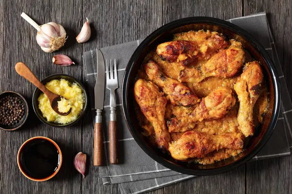 chicken drumsticks in crushed pineapple sauce in baking dish on dark rustic wooden table with ingredients and cutlery, horizontal view from above, flat lay