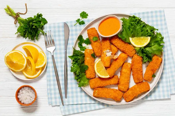 fried breaded fish sticks from white fish fillet served with fresh lettuce, lemon wedges and hot sauce on platter on white wooden table with cutlery, horizontal view from above, flat lay