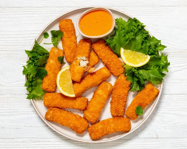 fried breaded fish sticks from white fish fillet served with fresh lettuce, lemon wedges and hot sauce on plate on white wooden table, horizontal view from above, flat lay, close-up