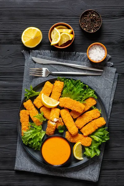 crispy fried breaded fish sticks from white fish fillet served with fresh lettuce lemon wedges and hot sauce on plate on black wooden table, vertical view from above