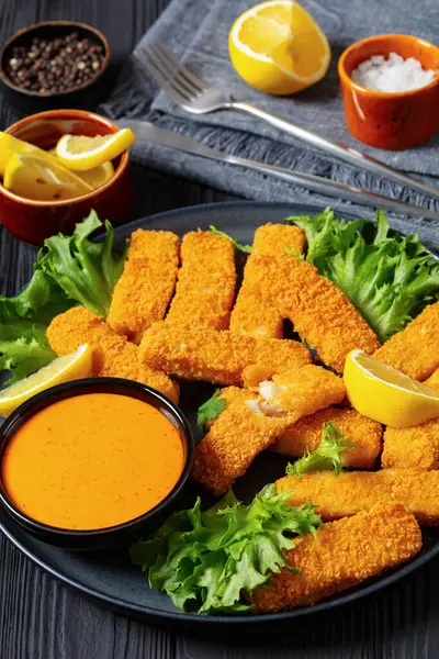 crispy fried breaded fish sticks from white fish fillet served with fresh lettuce lemon wedges and hot sauce on platter on black wooden table, vertical view, close-up