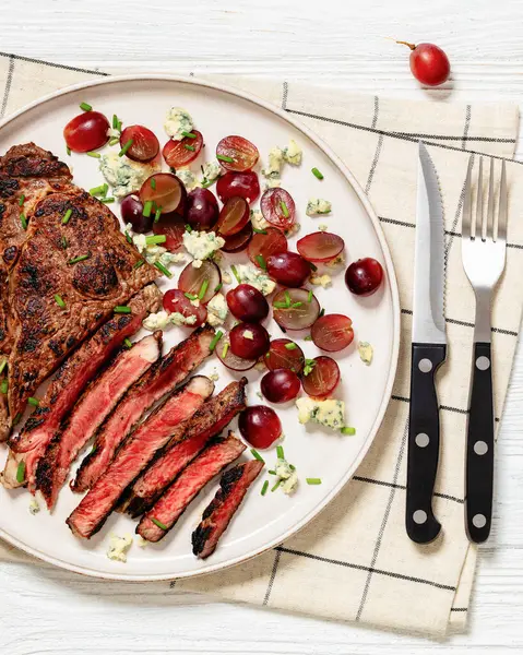 roasr beef steak served with salad of red grape, crumbled blue mold cheese and chives on plate on white wooden table with cutlery, horizontal view from above, flat lay, close-up
