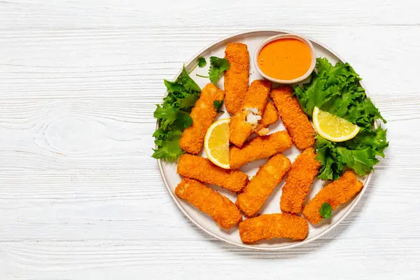fried breaded fish sticks from white fish fillet served with fresh lettuce, lemon wedges and hot sauce on plate on white wooden table, horizontal view from above, flat lay, free space