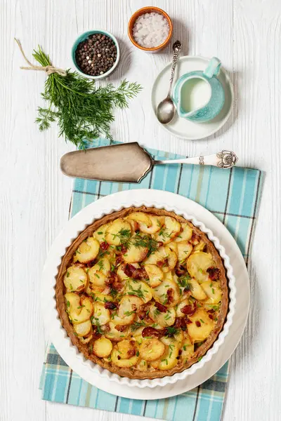 Irish potato pie of crispy crust layered with potatoes, bacon and onion in baking dish on white wooden table with ingredients and cake shovel, vertical view from above