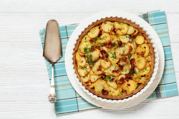 Irish potato pie of crispy crust layered with potatoes, bacon and onion in baking dish on white wooden table with cake shovel, horizontal view from above, flat lay, free space