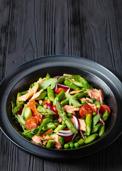 close-up of smoked salmon salad with asparagus, rocket salad, green peas, tomatoes and red onion in black bowl on black wooden table, vertical view, free space