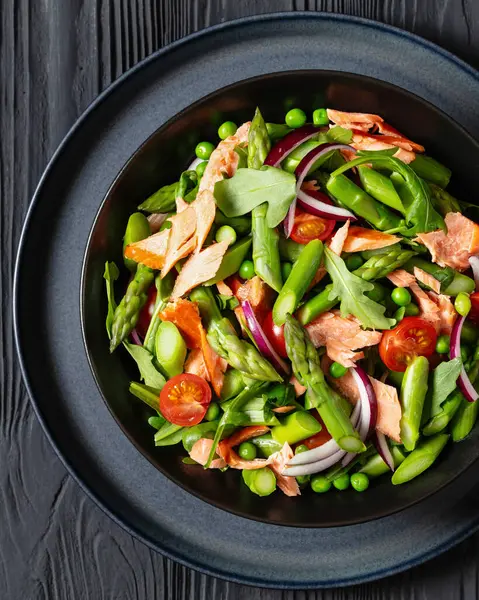 smoked salmon salad with asparagus, rocket salad, green peas, tomatoes and red onion in black bowl on black wooden table, flat lay, close-up, macro