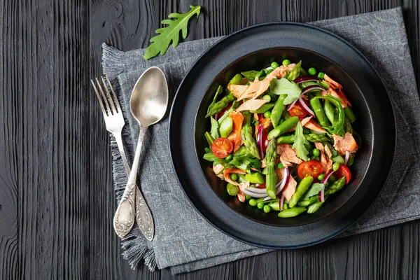 smoked salmon salad with asparagus, rocket salad, green peas, tomatoes and red onion in black bowl on black wooden table, overhead view, flat lay, free space