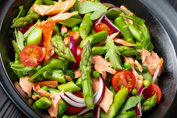 close-up of smoked salmon salad with asparagus, rocket salad, green peas, tomatoes and red onion in black bowl on black wooden table