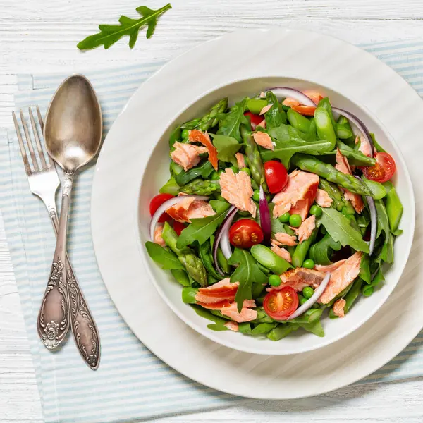 smoked salmon salad with asparagus, rocket salad, green peas, tomatoes and red onion in white bowl on white wooden table with fork and spoon, horizontal view from above, flat lay, close-up