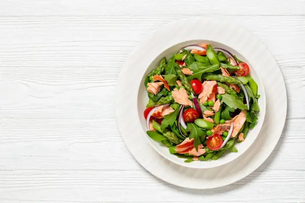 smoked salmon salad with asparagus, rocket salad, green peas, tomatoes and red onion in white bowl on white wooden table, horizontal view from above, flat lay, free space