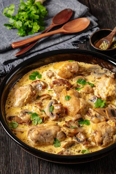 chicken thighs in a creamy mushroom garlic sauce with herbs and parmesan cheese in baking dish on dark wooden table with wooden spoons, vertical view, close-up