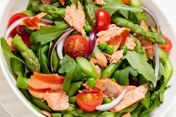 smoked salmon salad with asparagus, rocket salad, green peas, tomatoes and red onion in white bowl on white wooden table, close-up, dutch angle view