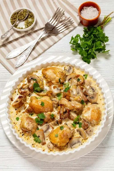 chicken thighs in a creamy mushroom garlic sauce with herbs and parmesan cheese in white baking dish on white wooden table with forks, vertical view from above