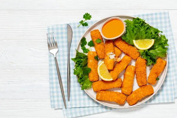 fried breaded fish sticks from white fish fillet served with fresh lettuce, lemon wedges and hot sauce on platter on white wooden table with cutlery, horizontal view from above, flat lay, free space