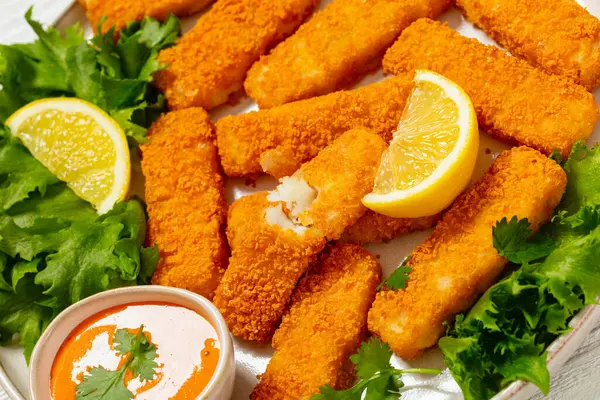 fried breaded fish sticks from white fish fillet served with fresh lettuce, lemon wedges and hot sauce on plate on white wooden table, close-up, dutch angle view