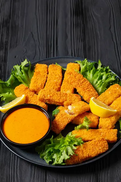 crispy fried breaded fish sticks from white fish fillet served with fresh lettuce lemon wedges and hot sauce on platter on black wooden table, vertical view, free space