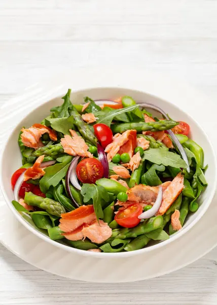 smoked salmon salad with asparagus, rocket salad, green peas, tomatoes and red onion in white bowl on white wooden table, vertical view from above, close-up