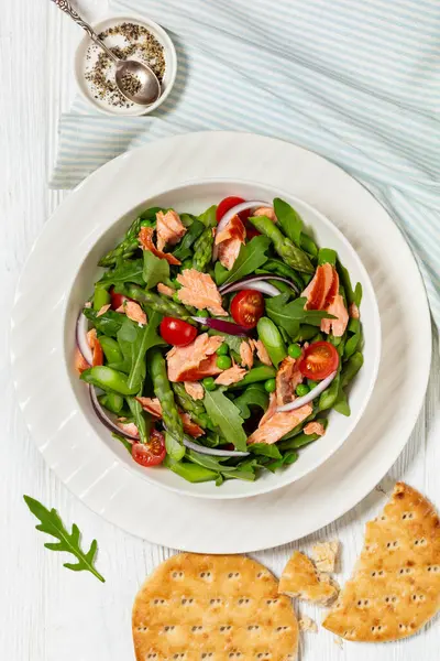 smoked salmon salad with asparagus, rocket salad, green peas, tomatoes and red onion in white bowl on white wooden table, vertical view from above