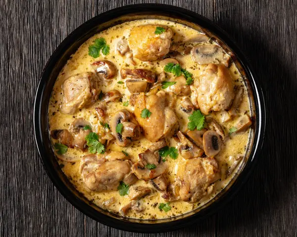 chicken thighs in a creamy mushroom garlic sauce with herbs and parmesan cheese in baking dish on dark wooden table, horizontal view from above, flat lay, close-up