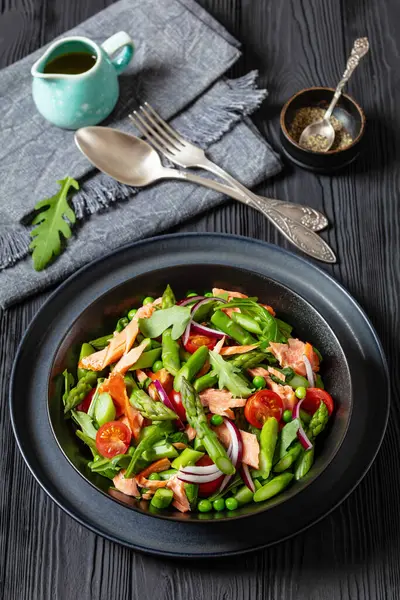 smoked salmon salad with asparagus, rocket salad, green peas, tomatoes and red onion in black bowl on black wooden table with fork and spoon, vertical view