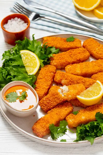 fried breaded fish sticks from white fish fillet served with fresh lettuce, lemon wedges and hot sauce on plate on white wooden table with cutlery, vertical view from above, close-up