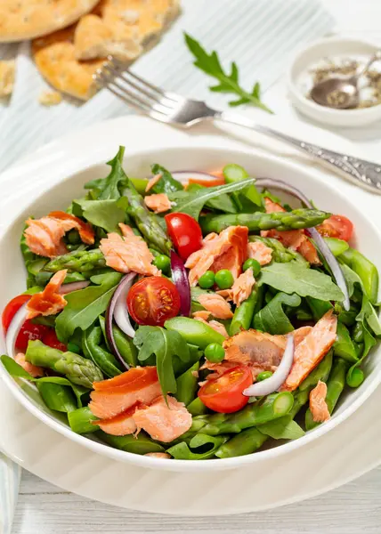 smoked salmon salad with asparagus, rocket salad, green peas, tomatoes and red onion in white bowl on white wooden table with flatbread, vertical view from above, close-up