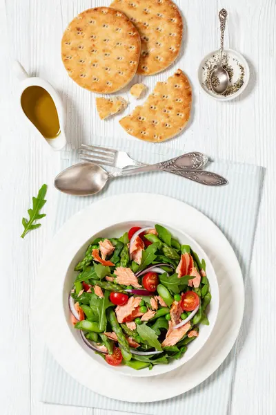 smoked salmon salad with asparagus, rocket salad, green peas, tomatoes and red onion in white bowl on white wooden table with flatbread, vertical view from above