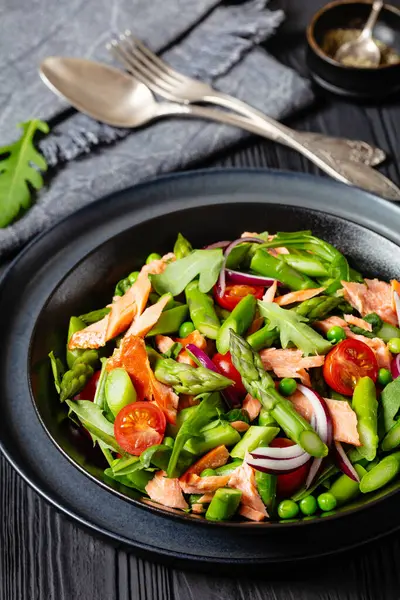 smoked salmon salad with asparagus, rocket salad, green peas, tomatoes and red onion in black bowl on black wooden table with fork and spoon, vertical view, close-up