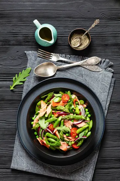 smoked salmon salad with asparagus, rocket salad, green peas, tomatoes and red onion in black bowl on black wooden table with fork and spoon, vertical view from above