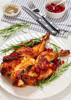 butterflied or spatchcock roast whole chicken on white plate with rosemary and cranberry on wooden table with cutlery, vertical view from above, close-up clipart