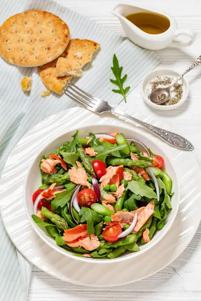 smoked salmon salad with asparagus, rocket salad, green peas, tomatoes and red onion in white bowl on white wooden table with flatbread olive oil, vertical view from above