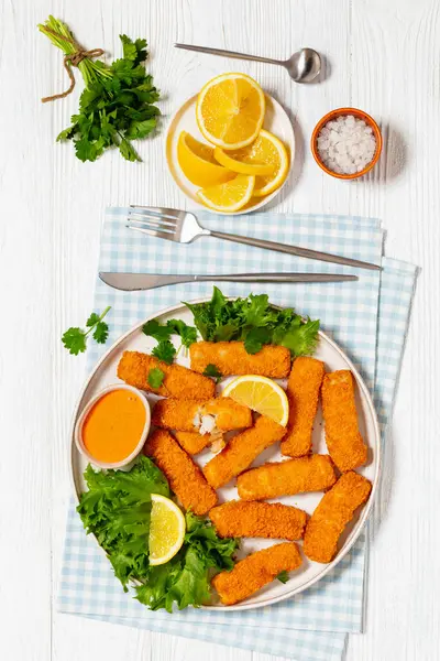 fried breaded fish sticks from white fish fillet served with fresh lettuce, lemon wedges and hot sauce on platter on white wooden table with cutlery, vertical view from above