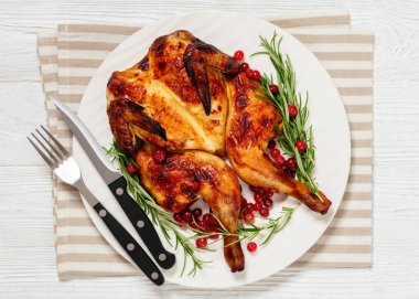 butterflied or spatchcock roast whole chicken on white plate with rosemary and cranberry on wooden rustic table with cutlery, horizontal view from above, flat lay, close-up clipart
