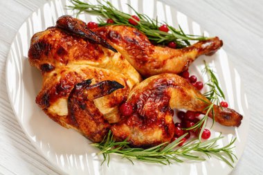 close-up of butterflied or spatchcock roast whole chicken on white plate with rosemary and cranberry on wooden table clipart