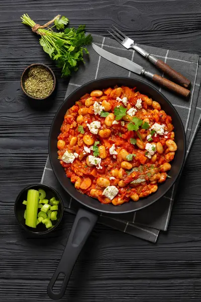 stock image gigantes plaki, greek giant baked beans in a chunky tomato sauce sprinkled with feta cheese and fresh cilantro on skillet on black wooden table with rustic fork and knife, vertical view