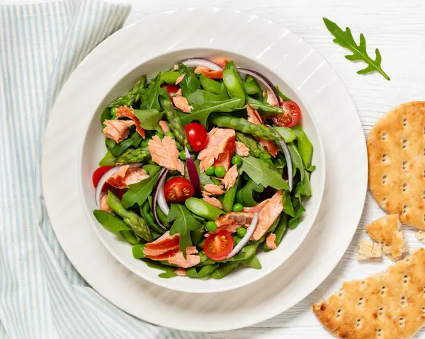 smoked salmon salad with asparagus, rocket salad, green peas, tomatoes and red onion in white bowl on white wooden table, horizontal view from above, flat lay,  close-up