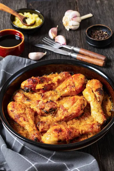 chicken drumsticks baked in crushed pineapple sauce in black dish on dark rustic wooden table with ingredients and cutlery at background, vertical view from above, close-up