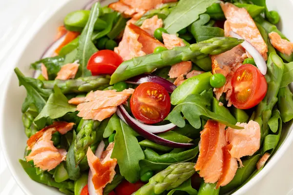 smoked salmon salad with asparagus, rocket salad, green peas, tomatoes and red onion in white bowl on white wooden table, close-up