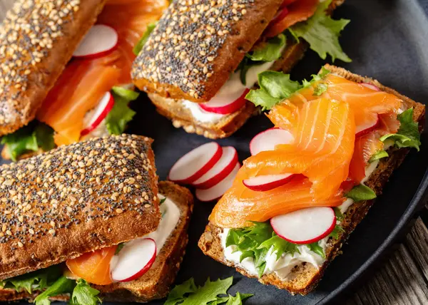 close-up of smoked salmon sandwiches on rye bread rolls with fresh red radish, lettuce and cream fresh on dark grey plate on rustic wooden table