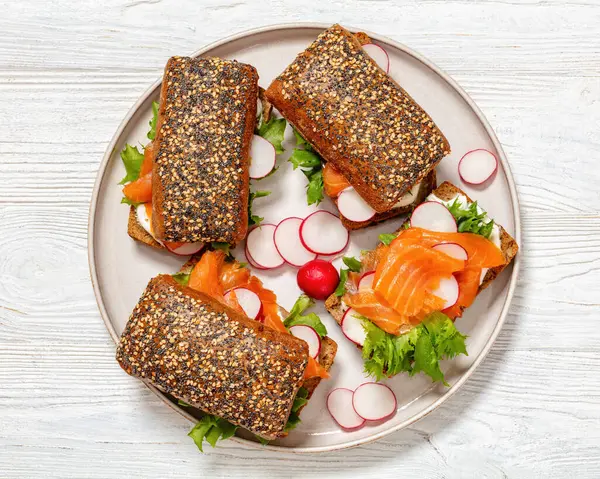 smoked salmon sandwiches on rye bread rolls with fresh red radish, lettuce and cream fresh on plate on white wooden table, horizontal view from above, flat lay,  close-up