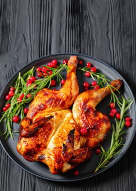 butterflied or spatchcock roast whole chicken on dark grey plate with rosemary and cranberry on black wooden table, vertical view, close-up clipart