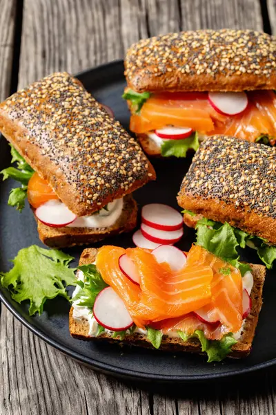 smoked salmon sandwiches on rye bread rolls with fresh red radish, lettuce and cream fresh on dark grey plate on rustic wooden table, vertical view, close-up