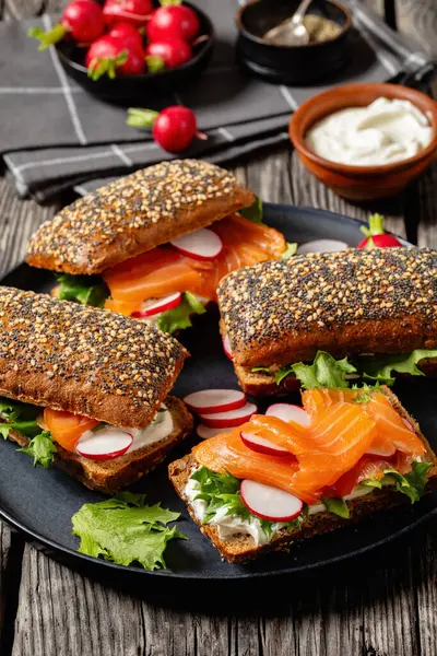 smoked salmon sandwiches on rye bread rolls with fresh red radish, lettuce and cream fresh on dark grey plate on rustic wooden table with ingredients, vertical view, close-up