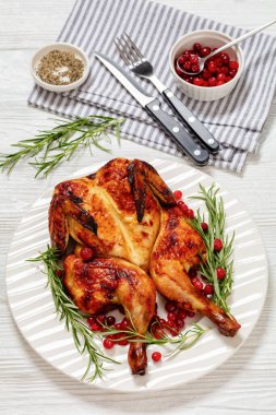 butterflied or spatchcock roast whole chicken on white plate with rosemary and cranberry on wooden table with cutlery and ingredients, vertical view from above clipart